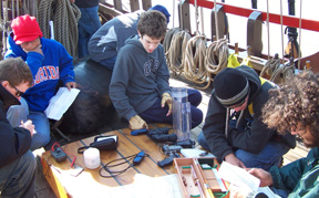 Students and adults sit around a collection of scientific instruments on the weather deck.