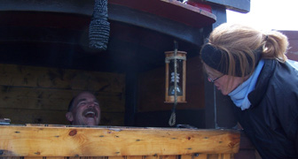 Captain Reynolds laughs while manning the helm and talking to Ms. Farrell.