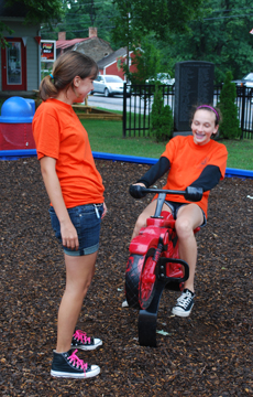 Evi cheers Nora on as the latter revs a playground motorcycle.